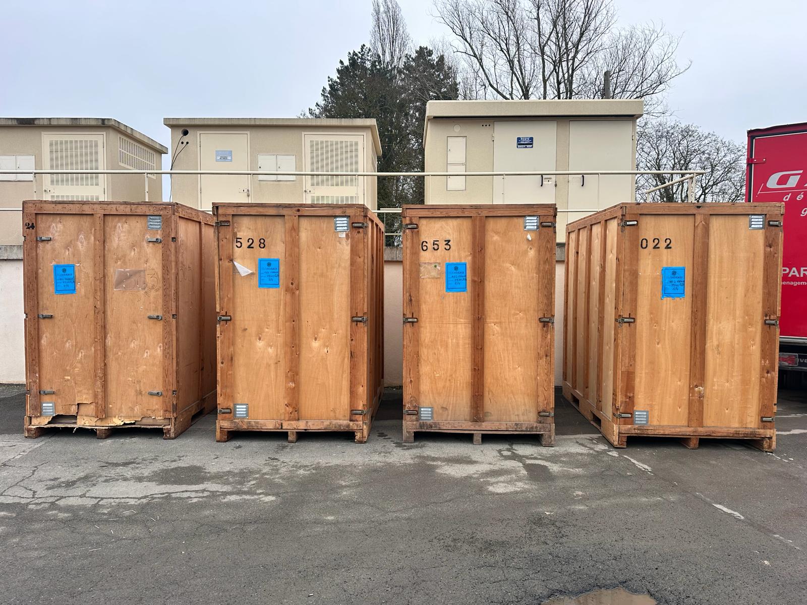 Quatre caisses de garde-meubles en bois numérotées et scellées, entreposées dans un centre de stockage sécurisé à Paris.