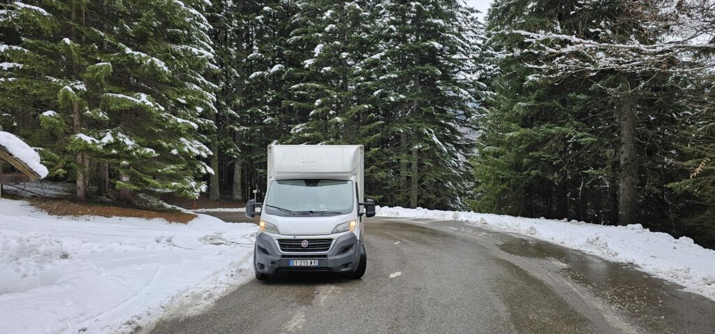 Camionnette de déménagement blanche garée sur un virage de route enneigée au cœur d'une forêt dense de sapins.