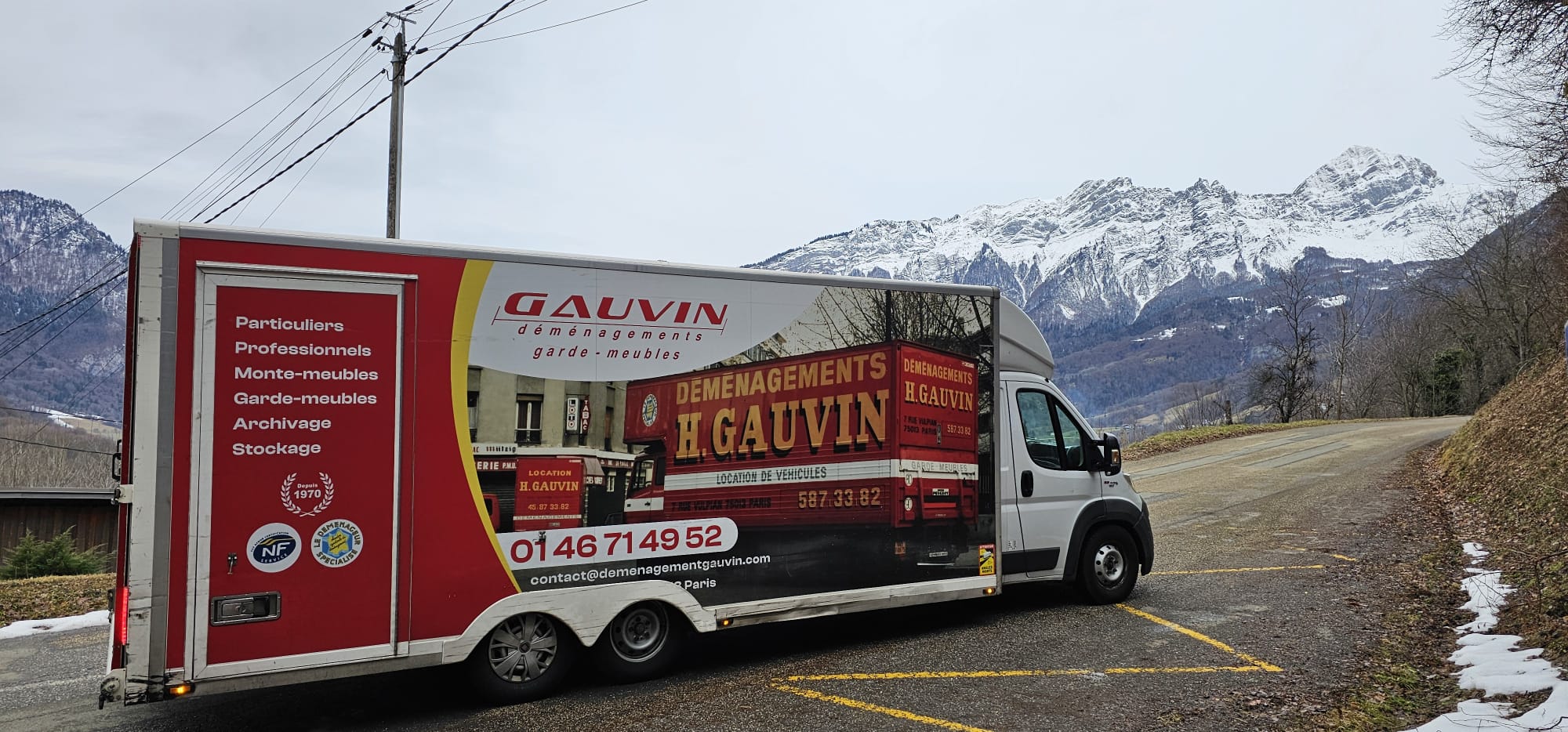 Camion de déménagement H. Gauvin rouge et blanc stationné le long d'une route avec de grandes montagnes enneigées en arrière-plan.