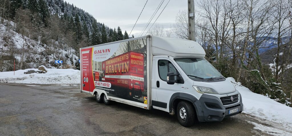 Camion utilitaire blanc et rouge floqué Déménagements H. Gauvin stationné sur le bas-côté d'une route de montagne enneigée.
