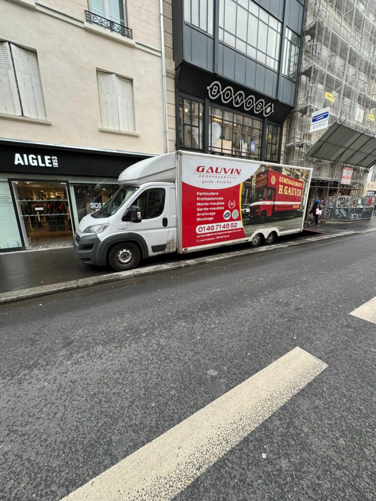 Camion utilitaire blanc et rouge des Déménagements H. Gauvin stationné le long d'une rue parisienne devant des commerces pour un chargement en ville.